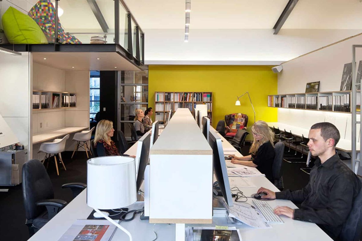 Office Layout Ideas: How to Plan a Better Workplace 1 Long shared bench desks in an open plan office with a yellow feature wall and bookshelves